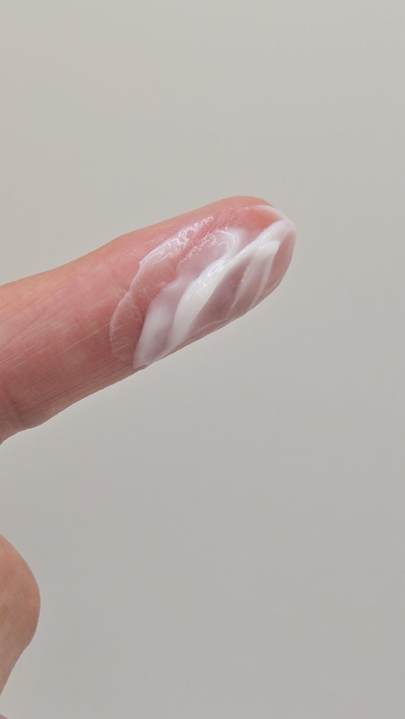 Puede incluir: Primer plano de un dedo con una porci&oacute;n de crema blanca. La crema tiene un aspecto liso, ligeramente texturizado, con crestas visibles y un brillo brillante. El fondo es gris claro liso.