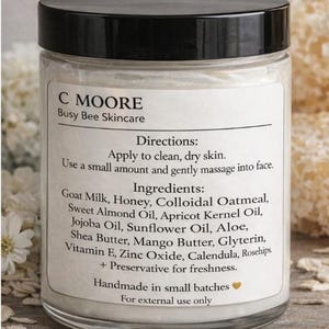 Puede incluir: Un tarro de vidrio transparente con tapa negra, etiquetado "C MOORE Busy Bee Skincare". La etiqueta enumera ingredientes como leche de cabra, miel y manteca de karit&eacute;. El tarro est&aacute; lleno de una crema blanca.