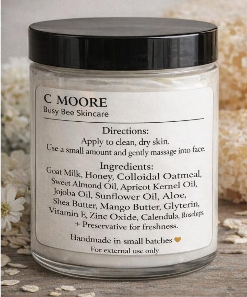 Puede incluir: Un tarro de vidrio transparente con tapa negra, etiquetado "C MOORE Busy Bee Skincare". La etiqueta enumera ingredientes como leche de cabra, miel y manteca de karit&eacute;. El tarro est&aacute; lleno de una crema blanca.
