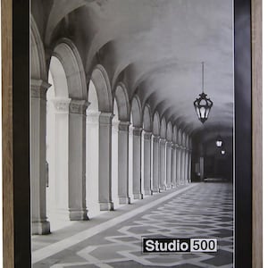 May include: A rectangular picture frame with a light brown wood-grain finish and a wide black mat border. The frame contains a black and white photograph of a long arched corridor with hanging lanterns. A Studio 500 logo is printed in the corner.