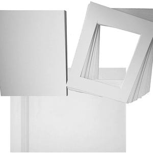 Puede incluir: Una pila de paspartús rectangulares blancos con bordes interiores biselados y una pila correspondiente de cartones de respaldo blancos. Los paspartús cuentan con un recorte rectangular central para enmarcar obras de arte o fotografías.