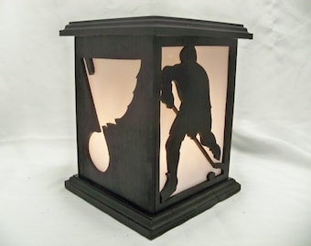 May include: A black wooden lamp with a silhouette of a hockey player and a hockey stick. The lamp has a white light inside and a white background.
