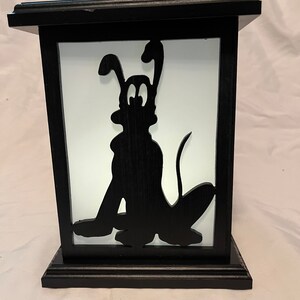 May include: Black wooden lantern featuring a silhouette of a cartoon dog. The dog is sitting with its tongue out. The lantern has a white background and a rectangular shape. The base and top of the lantern are also black.