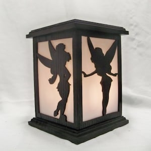 May include: A wooden lamp with a silhouette of two fairies on the front. The lamp has a white frosted glass panel and a dark brown wood frame.