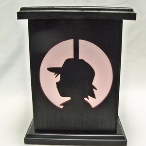May include: A black wooden box with a pink circle cutout featuring a silhouette of a person wearing a hat. The box has a raised edge around the top and bottom.