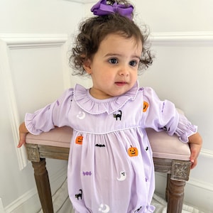 May include: A light purple baby romper with long sleeves and a ruffled collar. The romper is decorated with embroidered Halloween-themed images, including pumpkins, bats, ghosts, and cats. A large purple bow adorns the baby's head.