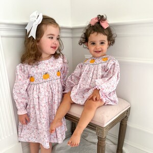 May include: Two young girls wearing pink floral smocked dresses with embroidered pumpkins. The girls are standing in a white room with white trim. One girl is sitting on a wooden stool with a pink cushion.