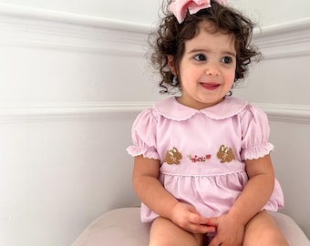 May include: A little girl wearing a pink dress with embroidered bunnies and flowers. She has curly brown hair and is wearing a pink bow.