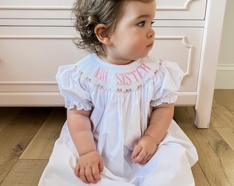 May include: A white smocked dress with pink embroidery and the words "Big Sister" embroidered on the neckline. The dress has a ruffled collar and short sleeves. A little girl is wearing the dress and has a pink bow in her hair.