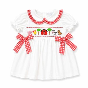 May include: A white short-sleeved dress featuring a red and white gingham Peter Pan collar and two matching side bows. The bodice displays smocking with embroidered farm animals, trees, and a red barn. The sleeves are gathered with ruffles.