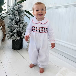 May include: A white baby romper with a red and white trim and a pattern of toy soldiers on the chest. The romper has long sleeves and a Peter Pan collar.