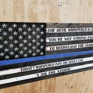 May include: A wooden American flag with a blue line, representing law enforcement, and a quote about strength and resilience. The quote reads: "The devil whispered in my ear "You're not strong enough to withstand the storm." Today I whispered into the devil's ear, "I am the storm."