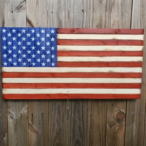 Puede incluir: Una bandera americana de madera pintada en rojo, blanco y azul. La bandera est&aacute; hecha de tablones de madera individuales y tiene un aspecto desgastado.