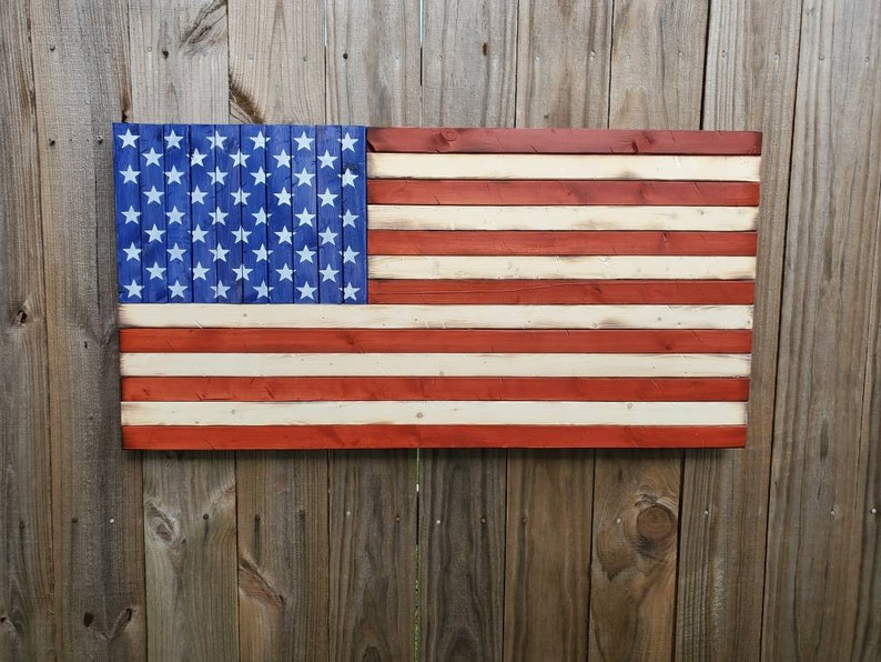 Puede incluir: Una bandera americana de madera pintada en rojo, blanco y azul. La bandera est&aacute; hecha de tablones de madera individuales y tiene un aspecto desgastado.