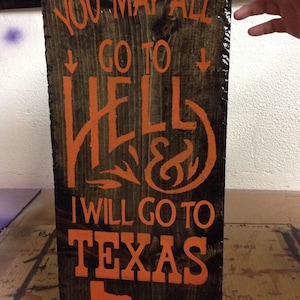 May include: A wooden sign with the text "You may all go to hell & I will go to Texas" in orange paint. The sign also features an outline of the state of Texas and the signature "Davy Crickett".