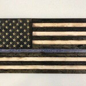 Op de afbeelding: Een Amerikaanse vlag van hout met een dunne blauwe lijn, die de steun voor de rechtshandhaving symboliseert. De vlag is gemaakt van gerecycled hout en heeft een verweerde afwerking. De sterren zijn van messing en zijn in het hout ingelegd.