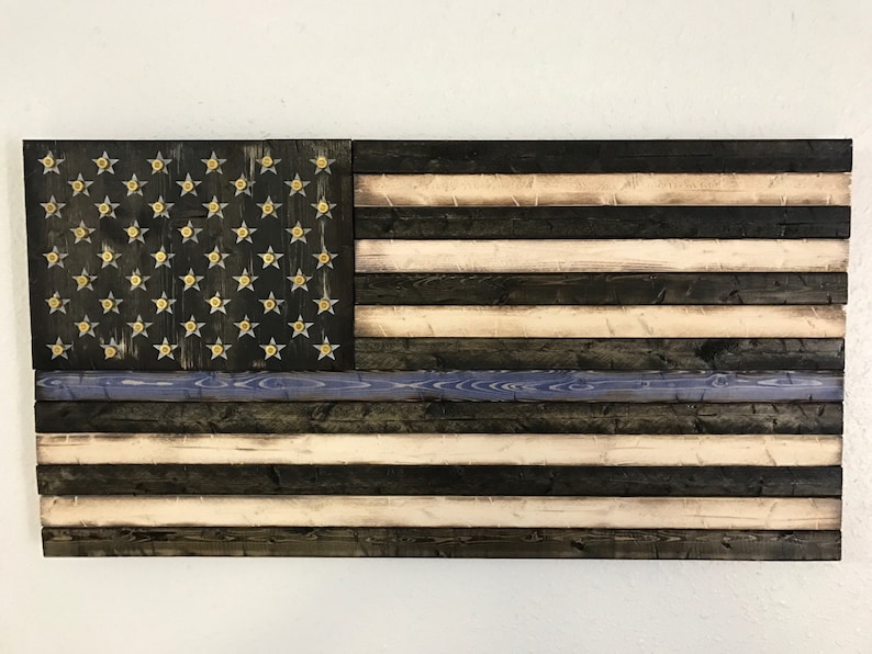 Op de afbeelding: Een Amerikaanse vlag van hout met een dunne blauwe lijn, die de steun voor de rechtshandhaving symboliseert. De vlag is gemaakt van gerecycled hout en heeft een verweerde afwerking. De sterren zijn van messing en zijn in het hout ingelegd.