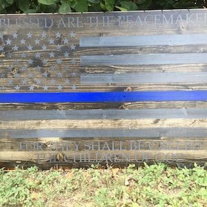 Może przedstawiać: Drewniana amerykańska flaga z niebieskim paskiem biegnącym poziomo przez środek. Flaga jest zużyta i ma postarzony wygląd. Tekst "Blessed are the peacemakers, for they shall be called the children of God" jest nadrukowany na dole flagi.