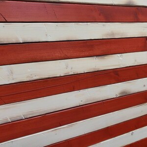 Puede incluir: Una bandera americana de madera r&uacute;stica con rayas rojas y blancas. La madera tiene un aspecto desgastado, lo que le da a la bandera un aspecto vintage.