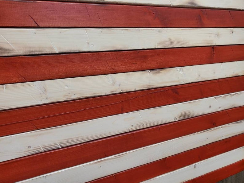 Puede incluir: Una bandera americana de madera r&uacute;stica con rayas rojas y blancas. La madera tiene un aspecto desgastado, lo que le da a la bandera un aspecto vintage.