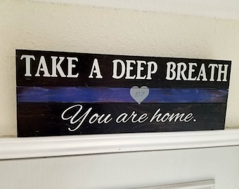 Könnte beinhalten: Ein rechteckiges Holzschild mit den Worten "TAKE A DEEP BREATH" und "You are home" in weißer Schrift. Eine blaue Linie und ein graues Herz mit "10-7" befinden sich in der Mitte. Das Schild ist aus dunkel gebeiztem Holz.