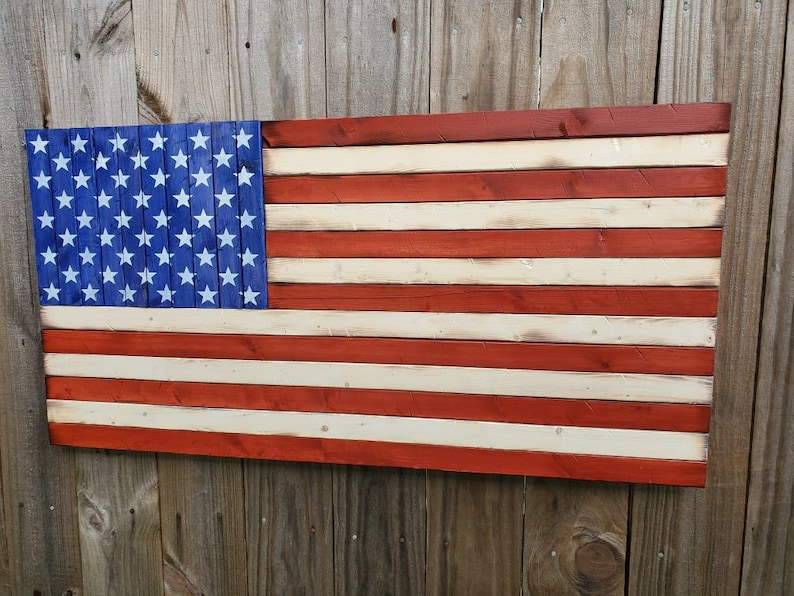 Puede incluir: Una bandera americana de madera pintada en rojo, blanco y azul. La bandera est&aacute; hecha de tablones de madera individuales, lo que le da un aspecto r&uacute;stico.