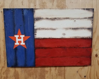May include: A wooden Texas flag with a red, white, and blue color scheme. The flag has a large orange star with a white "H" in the center, representing the Houston Astros baseball team.