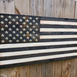 May include: A wooden American flag with a distressed black and white finish. The stars are made of gold-colored metal and are arranged in a traditional pattern. The flag is hanging on a wooden fence.