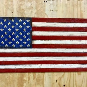 Op de afbeelding: Een Amerikaanse vlag van hout, geschilderd in rood, wit en blauw. De sterren zijn gemaakt van goudkleurig metaal en zijn bevestigd aan het blauwe veld. De vlag hangt aan een muur.