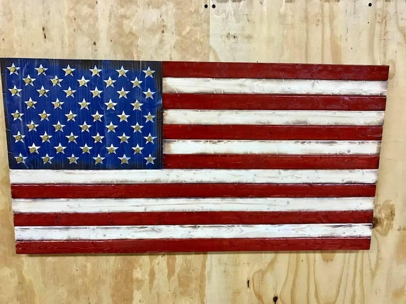 Op de afbeelding: Een Amerikaanse vlag van hout, geschilderd in rood, wit en blauw. De sterren zijn gemaakt van goudkleurig metaal en zijn bevestigd aan het blauwe veld. De vlag hangt aan een muur.