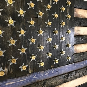 Op de afbeelding: Een Amerikaanse vlag van hout met een dunne blauwe lijn, met een verweerde zwarte achtergrond en gouden kogelhulzen voor de sterren.