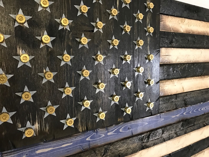 Op de afbeelding: Een Amerikaanse vlag van hout met een dunne blauwe lijn, met een verweerde zwarte achtergrond en gouden kogelhulzen voor de sterren.