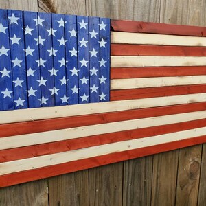 Puede incluir: Una bandera americana de madera pintada en rojo, blanco y azul. La bandera est&aacute; hecha de tablones de madera individuales y tiene un aspecto desgastado.