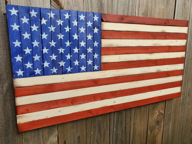 Puede incluir: Una bandera americana de madera pintada en rojo, blanco y azul. La bandera est&aacute; hecha de tablones de madera individuales y tiene un aspecto desgastado.