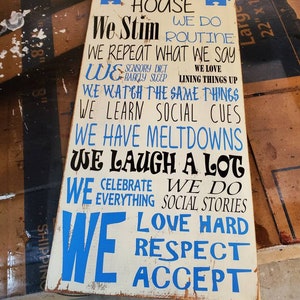K&ouml;nnte beinhalten: Ein wei&szlig;es Holzschild mit blauem Text, der lautet: "In diesem Haus stimulieren wir, wir machen Routine, wir wiederholen, was wir sagen, wir... lieben stark, respektieren, akzeptieren". Das Schild hat zwei blaue Puzzle-Teile in den oberen Ecken.