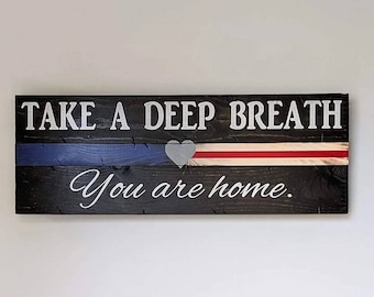 Op de afbeelding: Een houten bord met de tekst "Haal diep adem, je bent thuis." Het bord is zwart geverfd met witte letters en heeft een dunne blauwe lijn, een rode lijn en een wit hart.