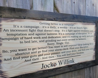 Könnte beinhalten: Ein Holzschild mit verbrannter Oberfläche zeigt ein motivierendes Zitat von Jocko Willink. Das Zitat lautet: "Besser werden ist eine Kampagne. Es ist eine Kampagne - es ist ein täglicher, ein wöchentlicher - es ist ein stündlicher Kampf. Ein unaufhörlicher Kampf, der nicht aufhört. Es ist ein Kampf gegen Schwäche und Versuchung und gegen Faulheit. Es ist eine Kampagne der Disziplin. Es ist eine Kampagne harter Arbeit und Hingabe, und es ist früh aufstehen und spät ins Bett gehen, und jede Sekunde dazwischen auskosten. Jeden. Einzelnen. Tag. Also, willst du besser werden? Willst du dich verbessern? Hör auf, nach einem Shortcut zu suchen und finde deinen Wecker. Finde deine Disziplin. Und finde deinen Mut und deine Leidenschaft und deine Motivation, und finde deinen Willen. Und dann - Und dann - wirst du deine Freiheit finden. Jocko Willink"