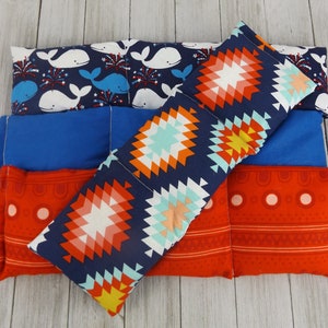 May include: Four fabric-covered heat packs in various patterns. The top pack has a navy blue background with white whales and red fireworks. The second pack is solid blue. The third pack has a navy blue background with a geometric pattern in red, orange, yellow, and turquoise. The bottom pack has a red background with a white geometric pattern.