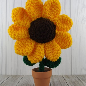 May include: A crocheted sunflower with yellow petals and a brown center, in a terracotta pot with green leaves.