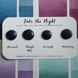 May include: Four black watercolour paint samples labelled "Stormfall", "Twilight Mist", "Evernight", and "Understory". The paint samples are arranged on a white card with the text "Into the Night" and a website address. The card is placed on a background of watercolour paper with various shades of blue, purple, and green.