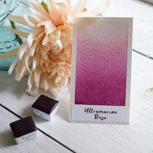 May include: A rectangular watercolor swatch card displaying a vertical gradient from pale pink to deep magenta, labeled with the text Ultramarine Rose. Two small square watercolor paint pans with dark purple pigment are positioned nearby.