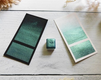 Op de afbeelding: Twee swatches van tealgroene metallic waterverf, een op zwart papier en een op wit papier, met een klein bakje van dezelfde verf.