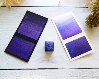 May include: Two watercolor swatches, one with a gradient from light to dark purple and the other a solid dark purple, and a small pan of dark purple watercolor paint.