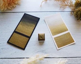 May include: Two swatches of gold watercolor paint, one on black paper and one on white paper, showing the color gradient from dark to light. A small pan of gold watercolor paint is in the center of the image.