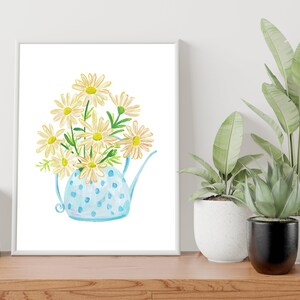 May include: A white framed print of a watercolor painting of a blue polka dot watering can filled with white daisies.