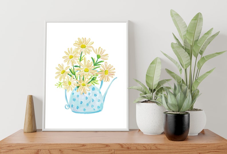 May include: A white framed print of a watercolor painting of a blue polka dot watering can filled with white daisies.