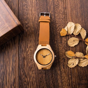 Puede incluir: Un reloj de pulsera de madera con una correa de cuero marr&oacute;n sobre un fondo de madera. La esfera del reloj es de color marr&oacute;n claro con manecillas y marcadores negros.