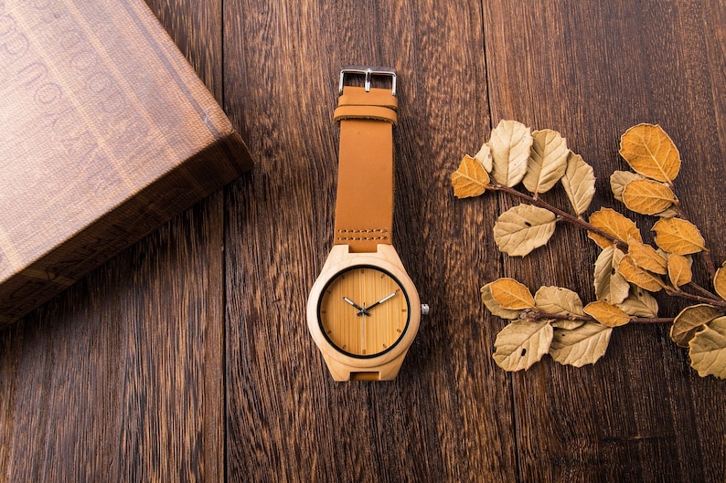 Puede incluir: Un reloj de pulsera de madera con una correa de cuero marr&oacute;n sobre un fondo de madera. La esfera del reloj es de color marr&oacute;n claro con manecillas y marcadores negros.