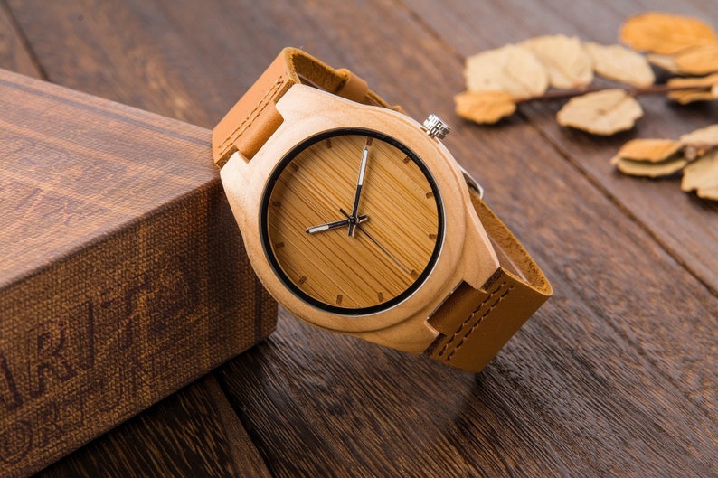 Puede incluir: Un reloj de pulsera de madera con una correa de cuero marr&oacute;n. La esfera del reloj es de madera y tiene agujas de hora y minuto negras.