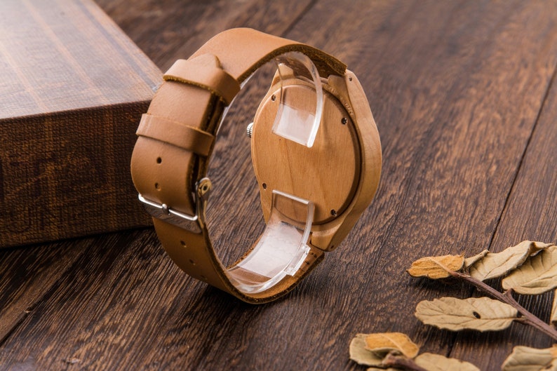 Puede incluir: Un reloj de pulsera de madera con una correa de cuero marr&oacute;n. La esfera del reloj es de madera y tiene un dise&ntilde;o sencillo.